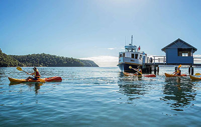 People kayaking on lake with ferry pulling into jetty with boatshed