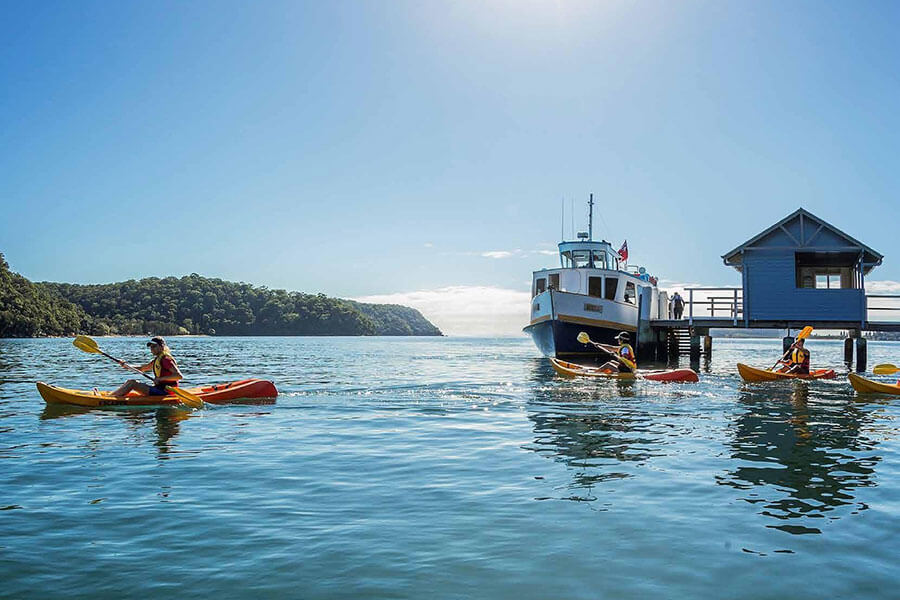 People kayaking on lake with ferry pulling into jetty with boatshed 