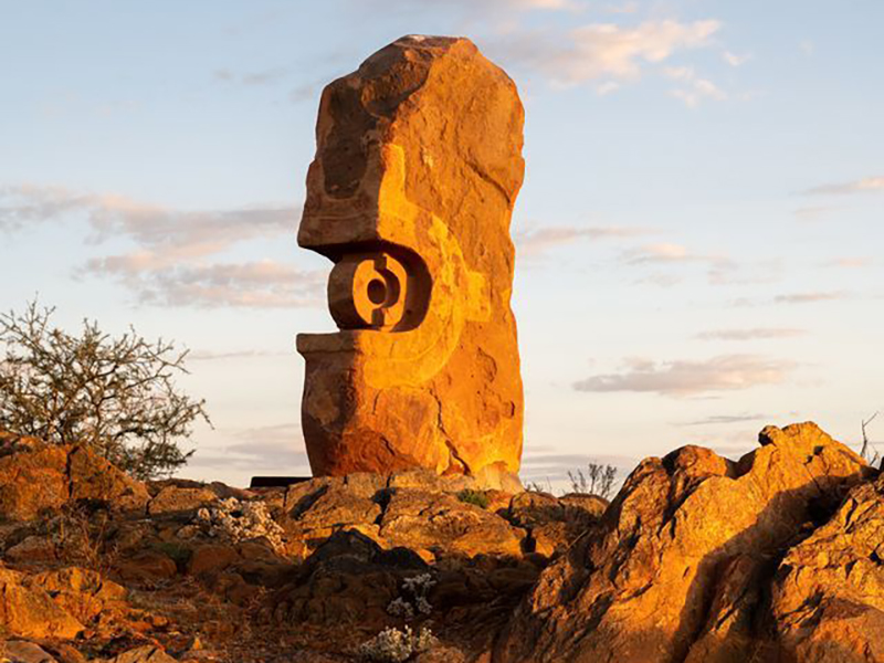 A tall stone statue in the desert. There are shrubs to one side of the statue and rocks to the other. It is sunrise or sunset and the light is reflecting off the statue, it looks golden and orange.