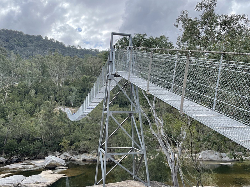 The new Bowtells swing bridge. The bridge is several metres off the ground above a shallow river. The bridge is metal, and connects two dirt tracks in the bush.