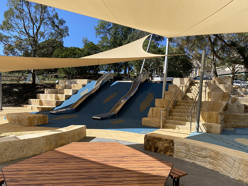 A shaded play area with two metal slides, sandstone stairs and seating and a timber picnic bench. There are trees and a lawn area in the background.