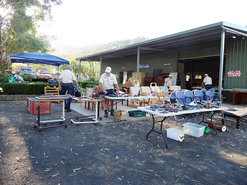 A large, corrugated iron building with a sign that says 'Men's Shed' on the front. There are people in matching yellow shirts setting up tables with tools and knick-knacks outside the building.