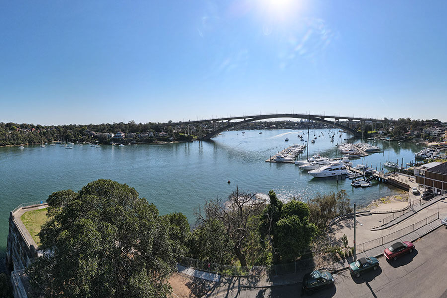 View overlooking water with boats, bridge in the background and cars parked on the side of the road.