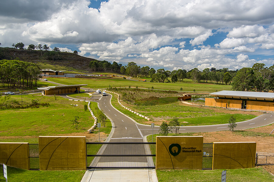 The new memorial site has a stone fence and metal gate, and a path from the entrance leading to two single-storey buildings. There is open greenspace and trees in the background.