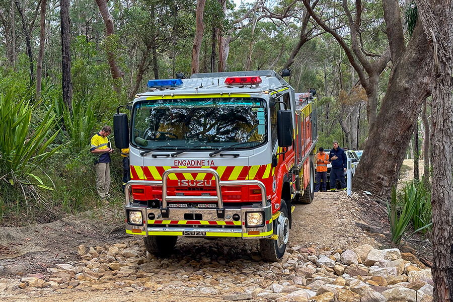 Fire truck on a dirt road in the bush. People are standing around the truck. There are trees and shrubbery in the background.
