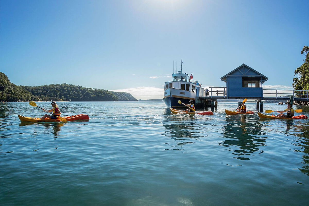 Friends kayaking on Pittwater as a ferry arrives at Bennetts Wharf, Coasters Retreat. Credit: Destination NSW